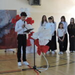 Uczeń szkoły podstawowej czyta przemówienie przy mikrofonie podczas uroczystości patriotycznej, a za nim stoi grupa uczniów oraz dekoracja z biało-czerwonymi kwiatami