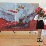 Uczennica gra na skrzypcach podczas występu artystycznego w szkolnej sali, na tle patriotycznej dekoracji z białym orłem