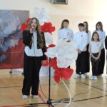 Uczennica śpiewa do mikrofonu na środku sali, a za nią stoi grupa młodzieży ubranej na galowo z przypiętymi biało-czerwonymi kokardami