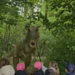 Dzieci stoją na leśnej ścieżce i obserwują realistyczny model tyranozaura w Parku Dinozaurów.