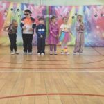 Przedszkolaki w kolorowych strojach inspirowanych latami 90. prezentują scenkę podczas występu z okazji Dnia Mamy na sali gimnastycznej.