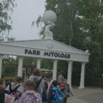Uczniowie spacerują w kierunku wejścia do Parku Mitologii. Nad bramą widoczna jest rzeźba postaci podtrzymującej kulę ziemską.