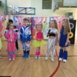 Grupa przedszkolaków w błyszczących, kolorowych kostiumach bierze udział w przedstawieniu inspirowanym muzyką i modą lat 90.