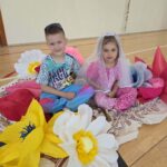 Dwoje dzieci w strojach scenicznych siedzi pośród dużych papierowych kwiatów przygotowanych jako dekoracja z okazji Dnia Mamy.