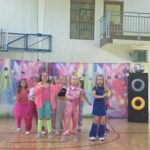 Przedszkolaki tańczą i prezentują układ choreograficzny podczas występu w stylu disco lat 90. na szkolnej sali gimnastycznej.