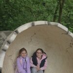Dwie dziewczynki siedzą w ogromnym modelu skorupy dinozaura przypominającej jajko. Wokół znajduje się zielony, leśny teren parku.