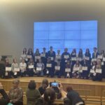 Na scenie sali konferencyjnej stoi grupa młodzieży trzymającej dyplomy i upominki podczas gali finałowej ogólnopolskiego konkursu „Auschwitz a postrzeganie współczesnego świata”. Uczniowie ubrani są odświętnie, a w tle widoczny jest duży ekran z nazwą wydarzenia. Przed sceną uczestników fotografują zgromadzeni goście i opiekunowie.