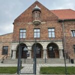 Ceglany budynek na terenie byłego obozu Auschwitz z charakterystycznym małym wykuszem nad wejściem. Budynek otoczony jest metalowym ogrodzeniem, a nad nim widoczne jest pochmurne niebo, podkreślające powagę miejsca pamięci.