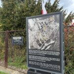 Tablica informacyjna na terenie byłego KL Auschwitz przedstawiająca archiwalne zdjęcie lotnicze żwirowiska oraz informacje dotyczące miejsca egzekucji więźniów obozu. Obok tablicy znajduje się ogrodzenie i tabliczka z napisem „Modlitwa w obronie Krzyża Klasztoru Sióstr Karmelitanek”.