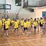 Uczennice prezentują występ taneczny na sali gimnastycznej. Dzieci w żółtych koszulkach poruszają się w zorganizowanym układzie choreograficznym.