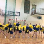 Grupa uczennic wykonuje układ taneczny z niebieskimi i żółtymi pomponami podczas występu artystycznego w ramach Dnia Sportu.