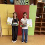 Dwie uczennice – laureatki konkursu „Olimpusek z matematyki” – stoją w sali lekcyjnej, trzymając dyplomy oraz nagrody książkowe i edukacyjne.