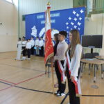 Poczet sztandarowy oraz uczniowie podczas uroczystości zakończenia roku szkolnego w sali gimnastycznej.