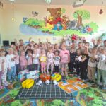 Grupa dzieci stoi wokół dużej maty do kodowania. Na macie znajdują się trzy roboty edukacyjne, pojemniki z kartami i elementami do programowania oraz plansza z zadaniami.