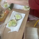 Na szkolnej ławce leżą kanapki z pełnoziarnistego pieczywa, posmarowane serkiem i udekorowane plasterkami ogórka, żółtym serem oraz sałatą. Obok znajdują się świeży ogórek, liście sałaty i opakowanie serka. W górnej części zdjęcia widoczne są dłonie sięgające po sałatę.