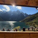 Widok na Morskie Oko i ośnieżone szczyty Tatr z tarasu schroniska. Nad jeziorem świeci słońce, a przy drewnianej barierce stoją turyści.